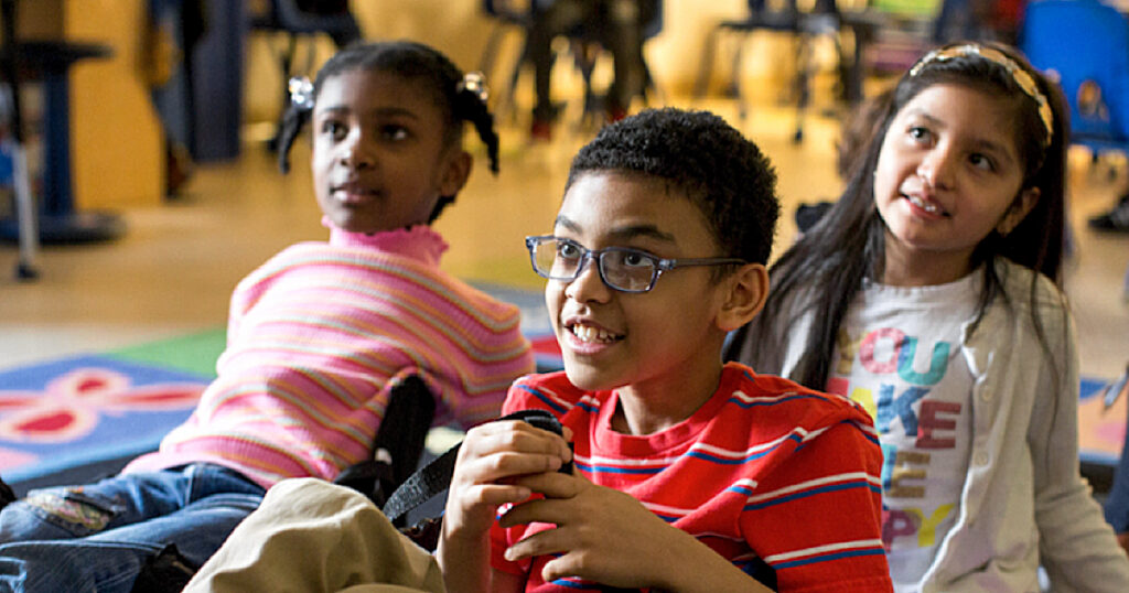 Three diverse children sitting on the floor of a classroom engaged in a lesson