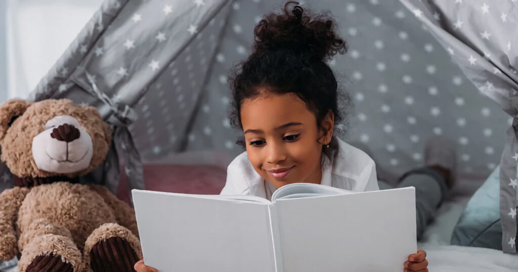 Young Black girl reading a book in front of a tent with a teddy bear.