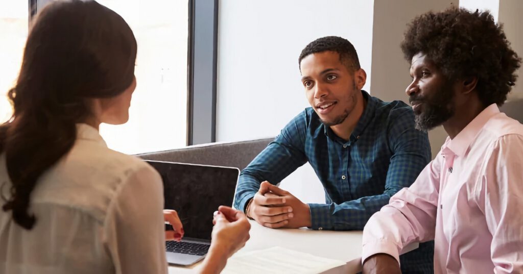 Black father and teenage son sitting at a table, meeting with a Black woman teacher for a conference.