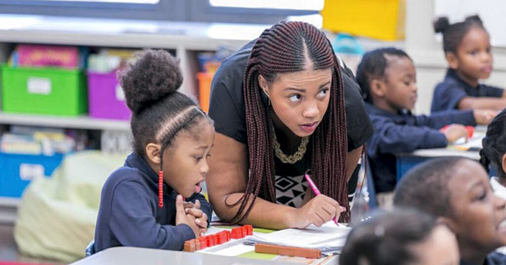African American woman leaning over a desk with a pencil teaching African American students sitting at their desks in a classroom at Uncommon Schools.