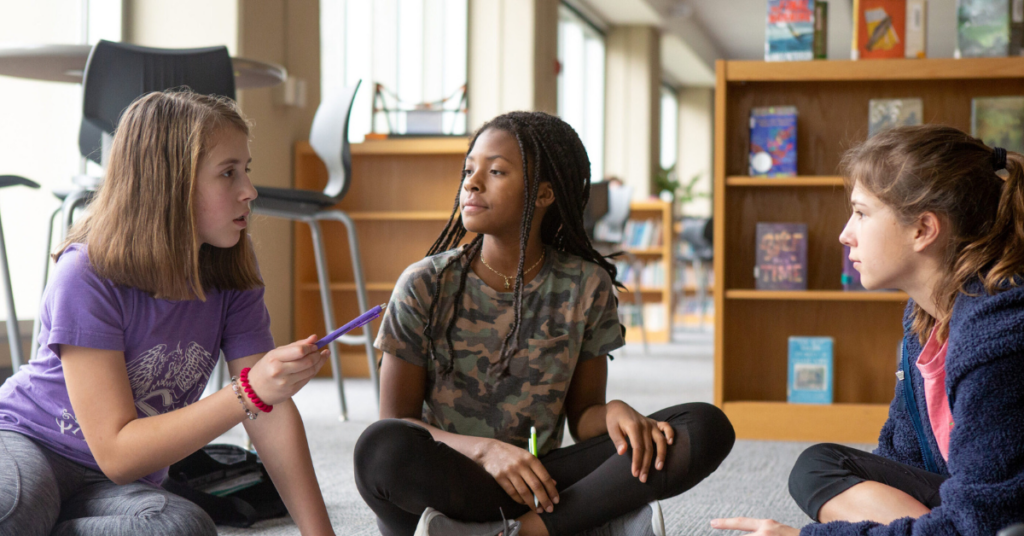 Two white female students and one Black female student sitting on the floor in a classroom.