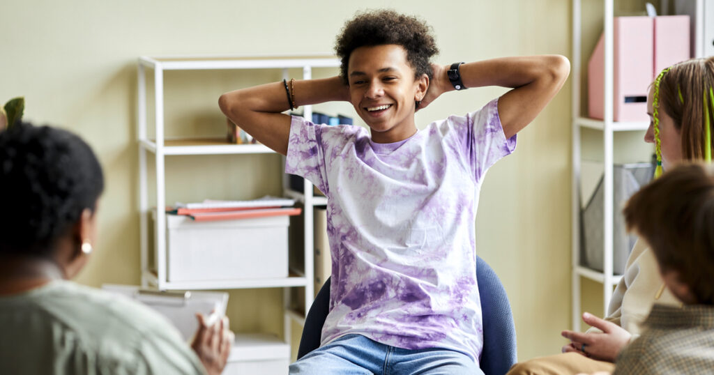 Young Black male student smiling with his hands behind his head, talking in a group session.