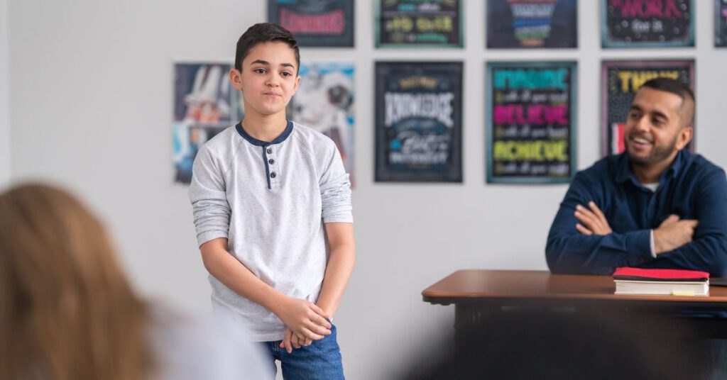 Young boy standing at the front of a classroom with his hands crossed in front of him