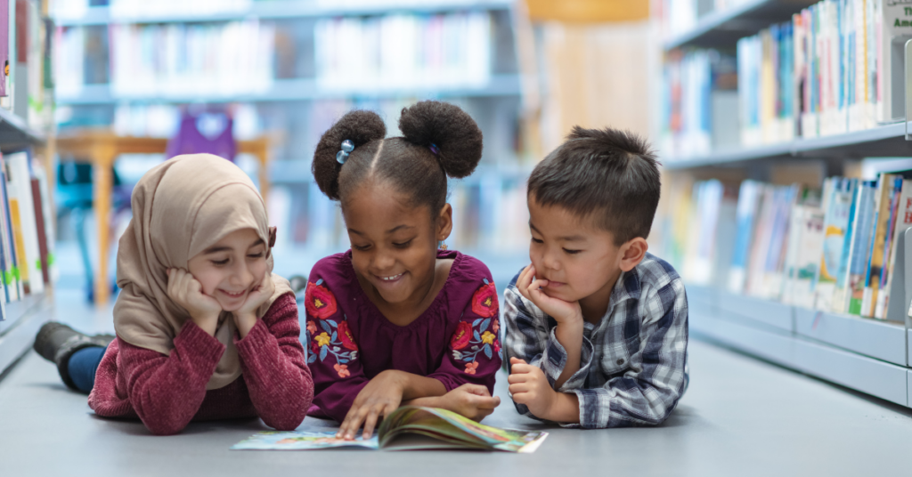 Three diverse children laying on a library floor reading a book together.