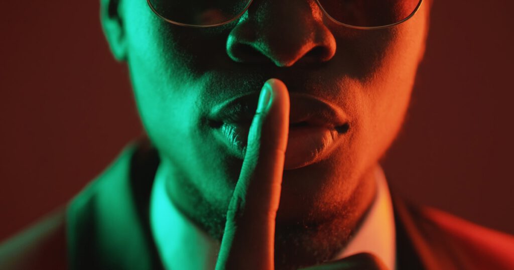 Shush gesture. Man secret. Confidential conspiracy. Green red neon light unrecognizable gentleman in suit glasses warning shh with finger on lips on dark.