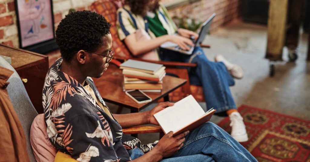 African American guy reading book while sitting in armchair against his Caucasian girlfriend typing on laptop keyboard in living room