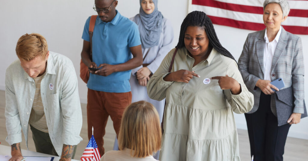 Multi-ethnic group of people registering at polling station decorated with American flags on election day, focus on smiling African woman pointing at I VOTE sticker, copy space