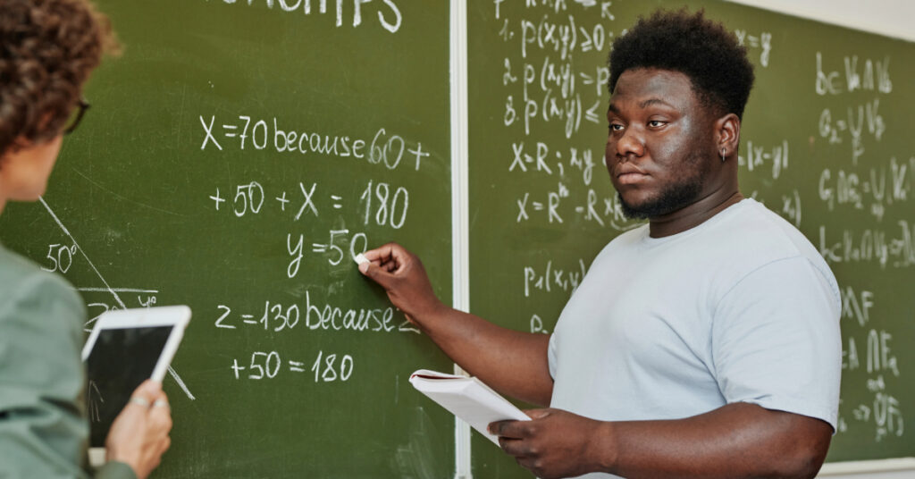 Youthful African American student pointing at blackboard during explanation of equation or theorem and looking at teacher