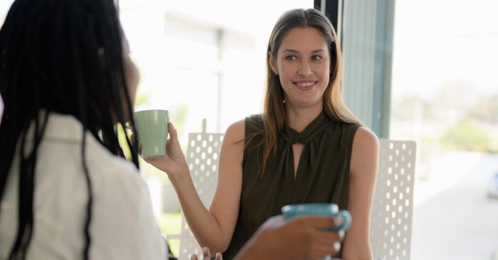 Happy white female talking with black woman while drinking coffee.