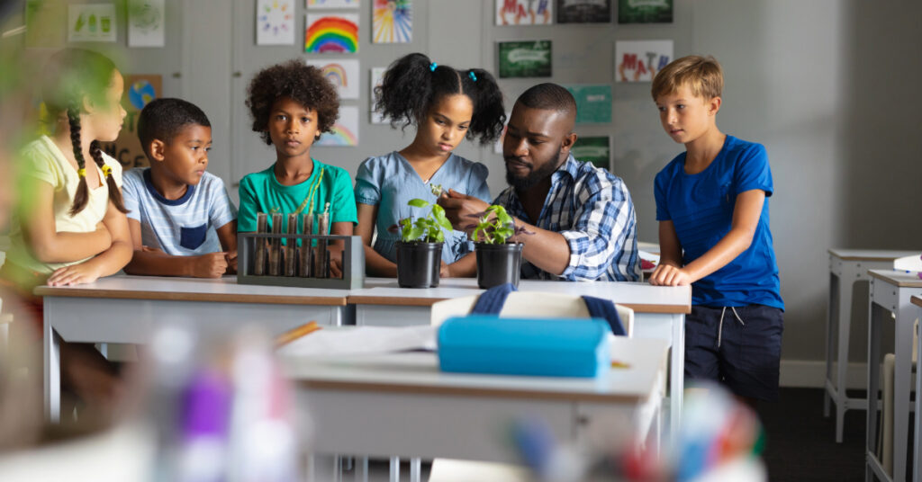 African american young male teacher showing plants to multiracial elementary students in class. unaltered, education, childhood, teaching, science, stem and school concept.