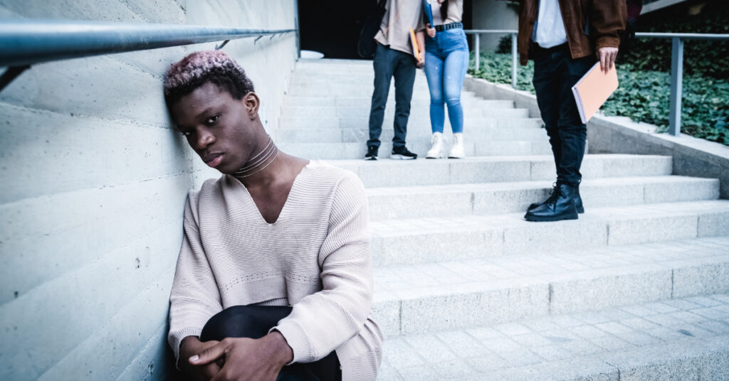 Upset African American transgender student leaning on wall near crop classmates throwing crumpled paper outside university building