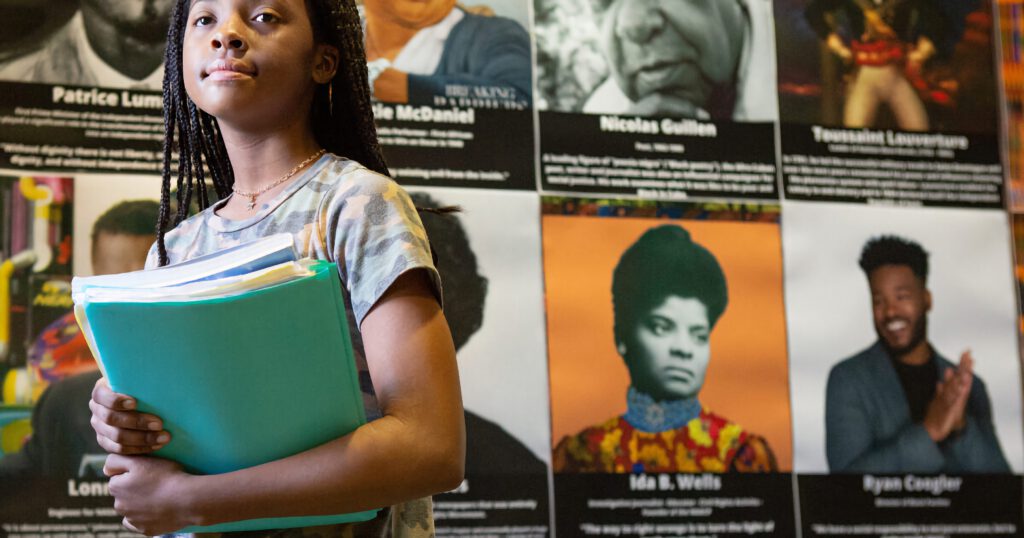 Young African American girl with braids holding a binder, standing in front of a wall of photos of important African Americans in History, Museum Exhibit or School Bulletin Board