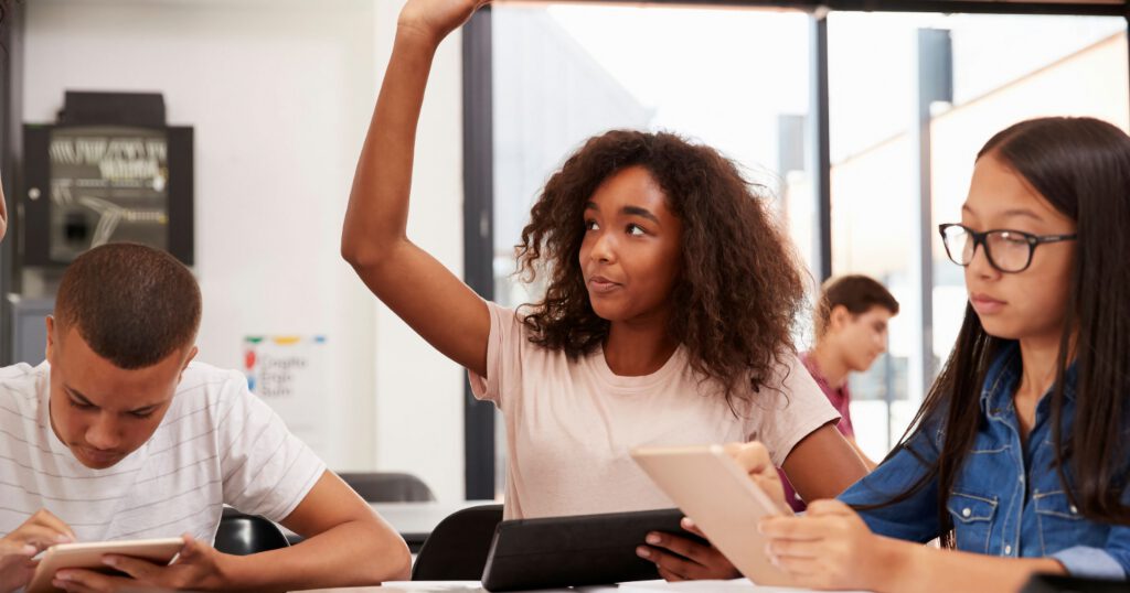 Diverse teenage students sitting at a table in a classroom, Black female student with her hand up waiting to ask a question