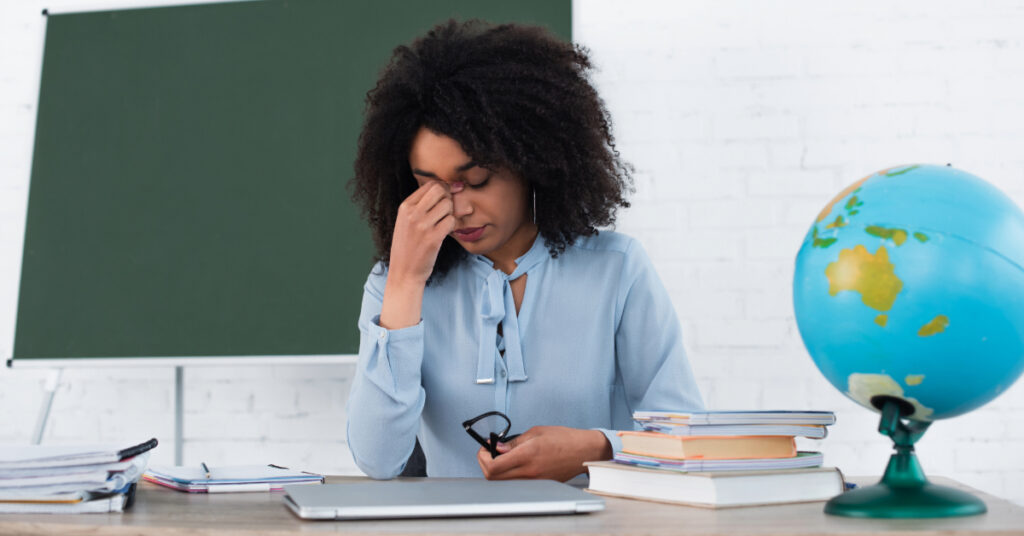 Tired african american teacher holding eyeglasses near laptop and globe in classroom