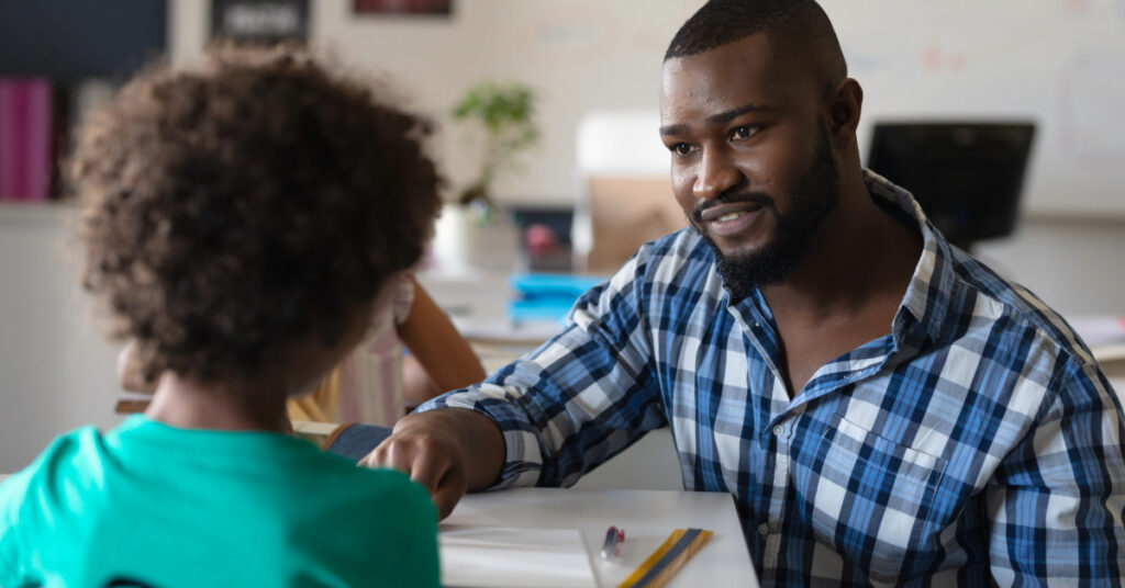 African american young male teacher talking on african american elementary schoolboy sitting at desk