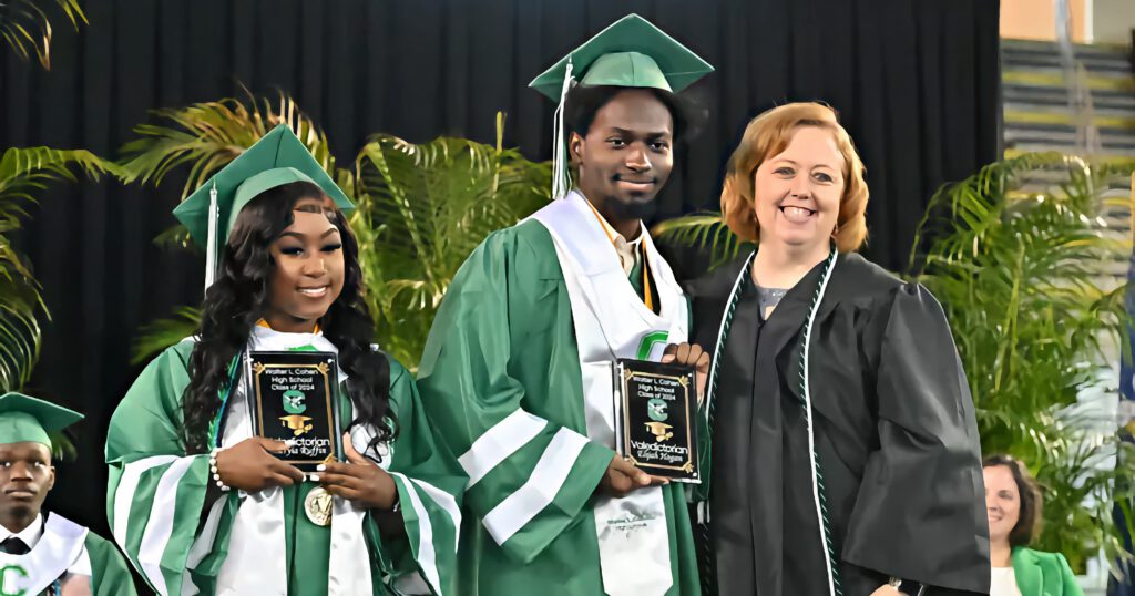 Elijah Hogan in green graduation regalia, accepts the honor of valedictorian at his high school graduation from a faculty member. Fellow graduates in the background.