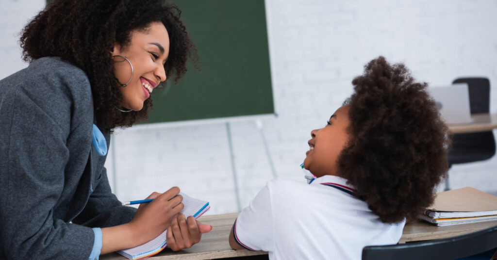 Smiling african american teacher writing on notebook near pupil in classroom