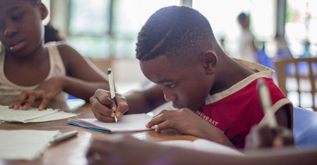 Young black male student works on classwork while seated at a table next to a black female student.