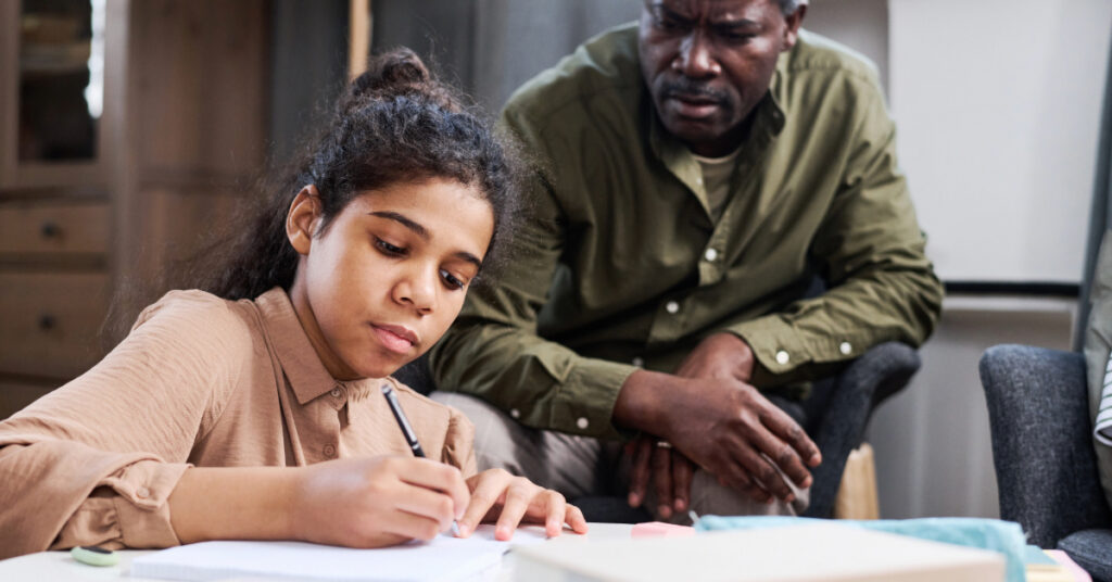 Serious multi-ethnic schoolgirl making notes in copybook while doing homework and her grandfather checking assignment