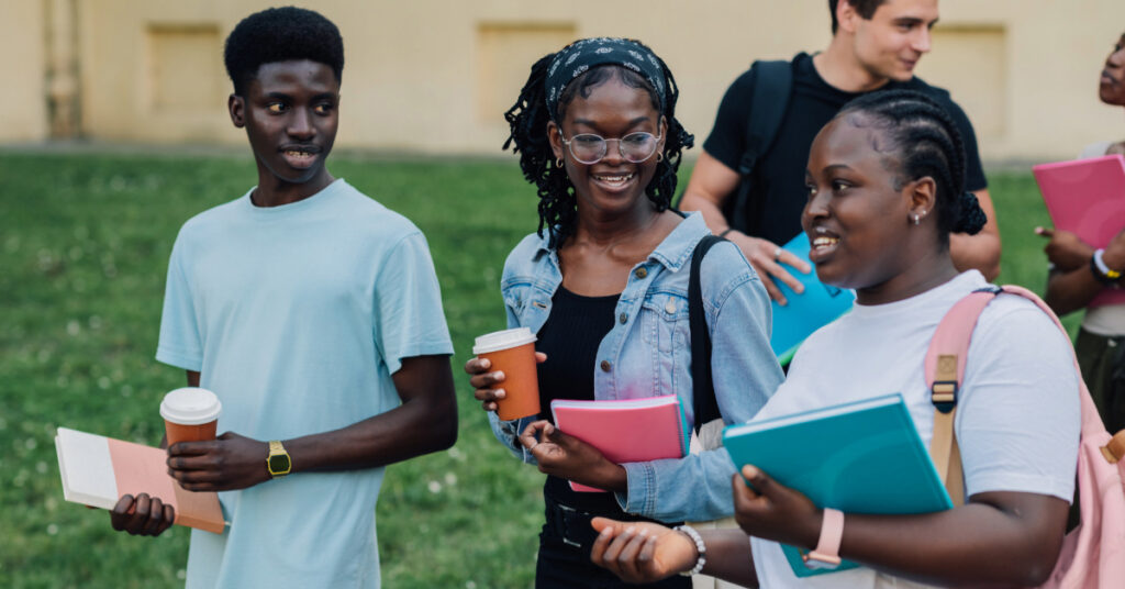 Portrait of small group of multicultural students with textbooks and takeaway coffees in hands walking in crowd near university building at campus, chatting and smiling at each other after lectures.