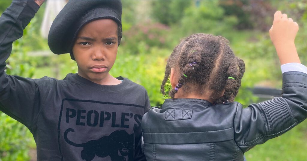 A young Black boy and girl dressed in black clothing, raising their fists.