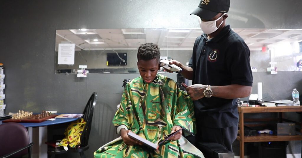 Young Black boy sitting in a barber chair reading a book while a Black barber cuts his hair. Mirror and chess set in the background.