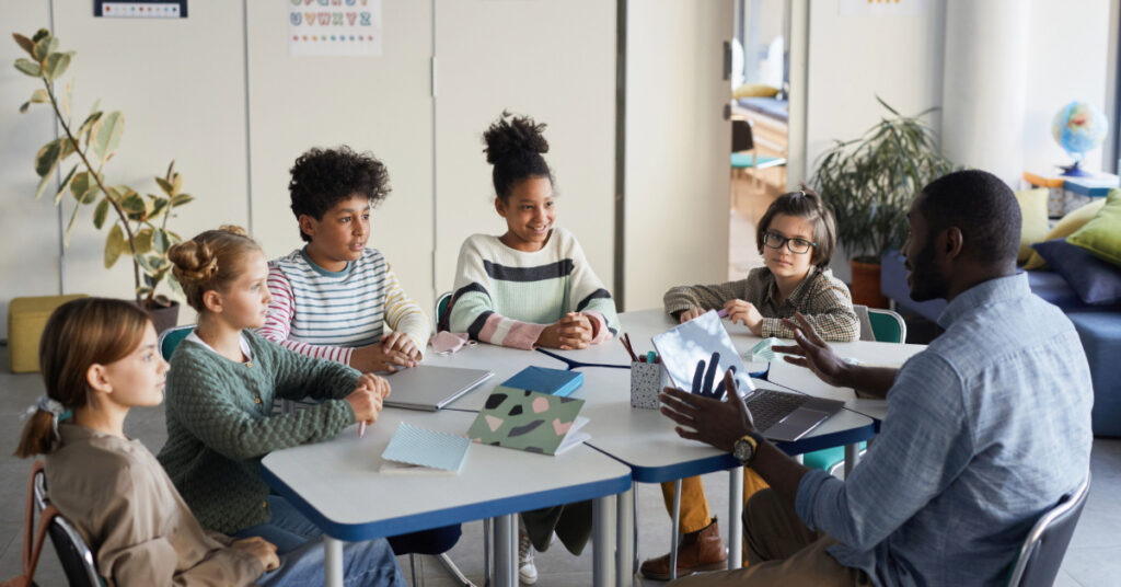 Diverse group of children sitting at table with male teacher in modern school classroom