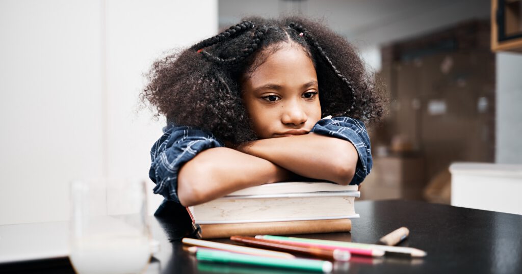Shot of a young Black girl laying her chin on her arms over a stack of books looking unhappy while doing a school assignment