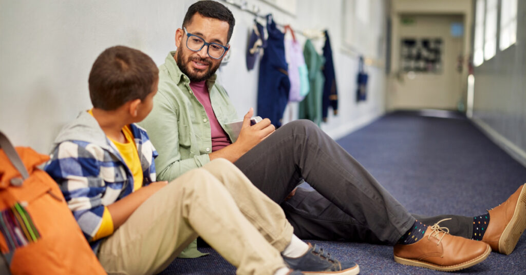 Young and friendly teacher speaking to boy in school hallway.