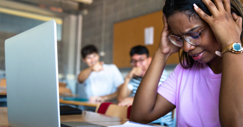 Sad female black teen high school student being bullied by classmates in class.
