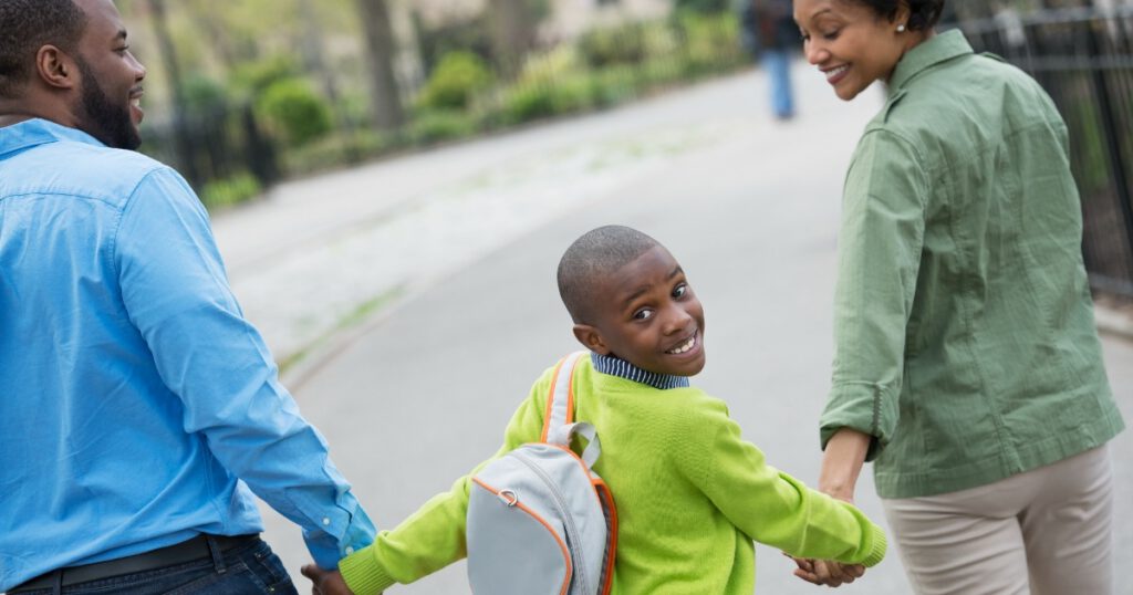A New York city park in the spring. A boy with a book bag, holding hands with his mother and father.