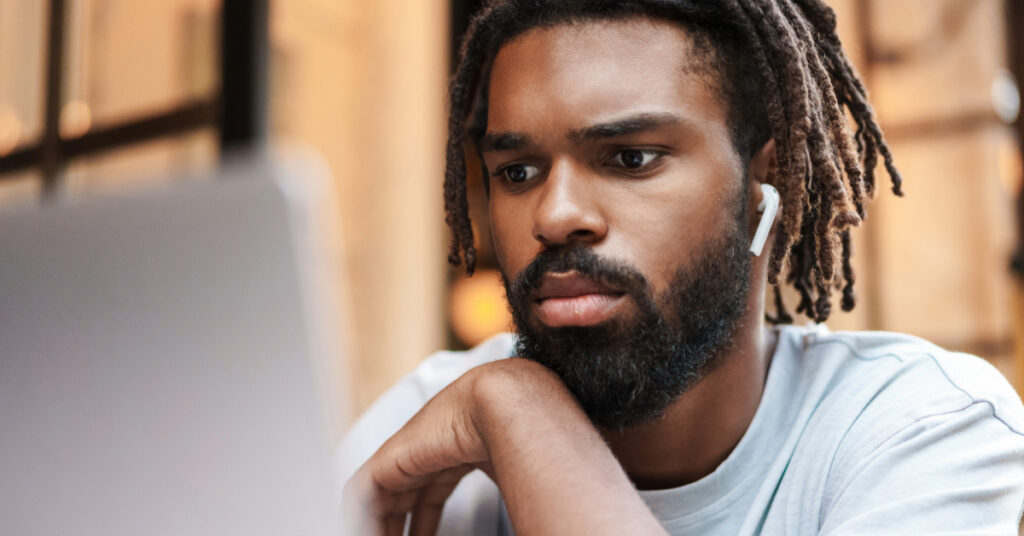 Focused african american guy in earphones working with laptop while sitting in cafe outdoors