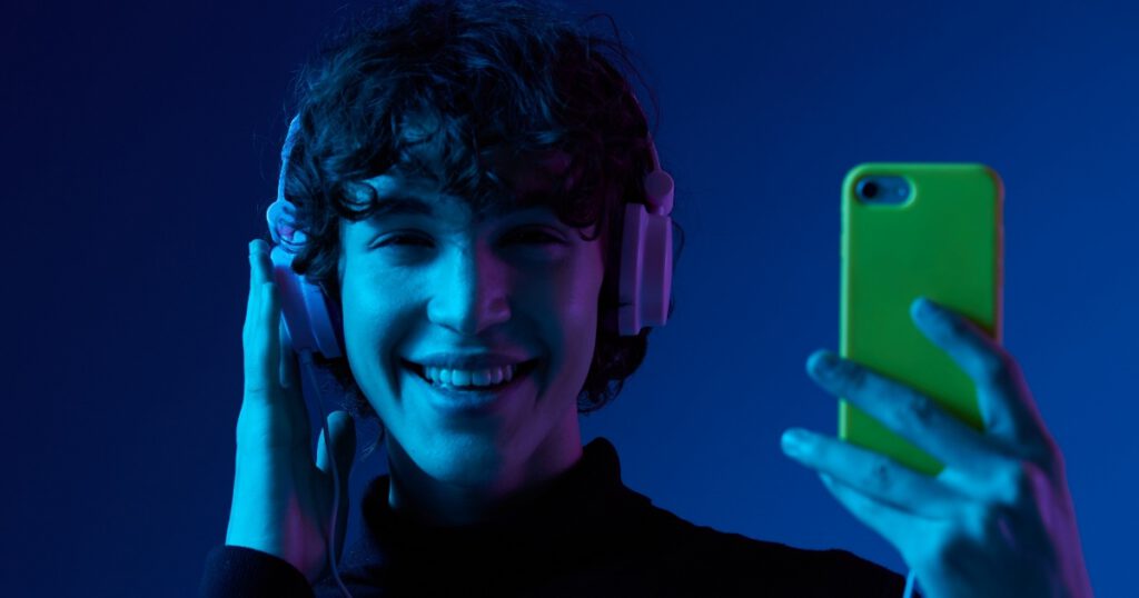 Young male in blue light wearing headphones holding a mobile phone and smiling