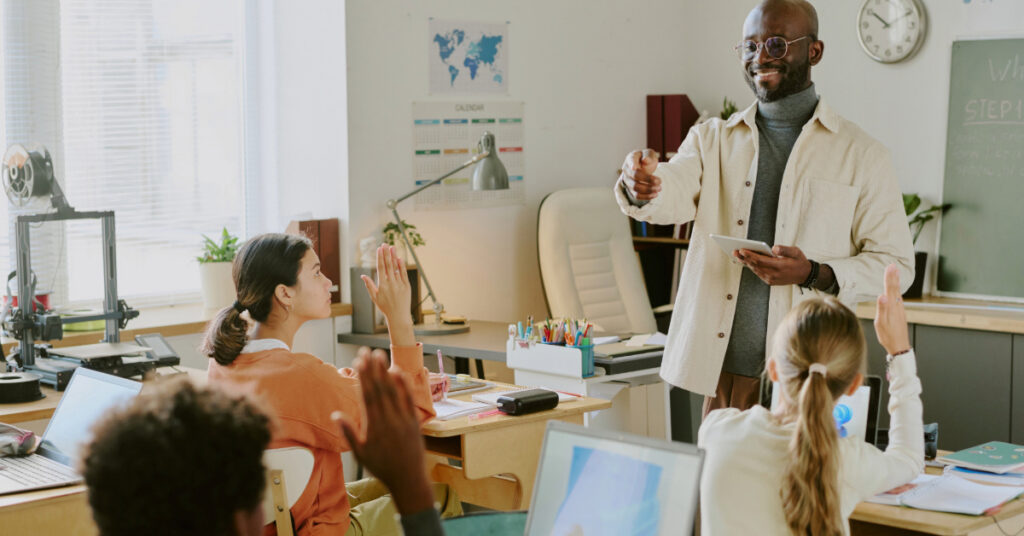 Educator explaining a concept in bright classroom while students attentively listen and raise hands.
