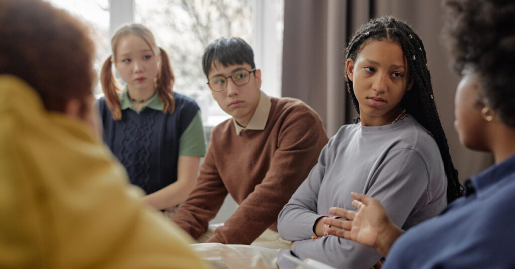 Multiethnic group of teenage children listening to female therapist during support session in school or community center