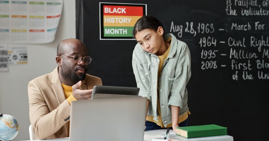 Black male teacher talking to his student in class with a Black History Month background