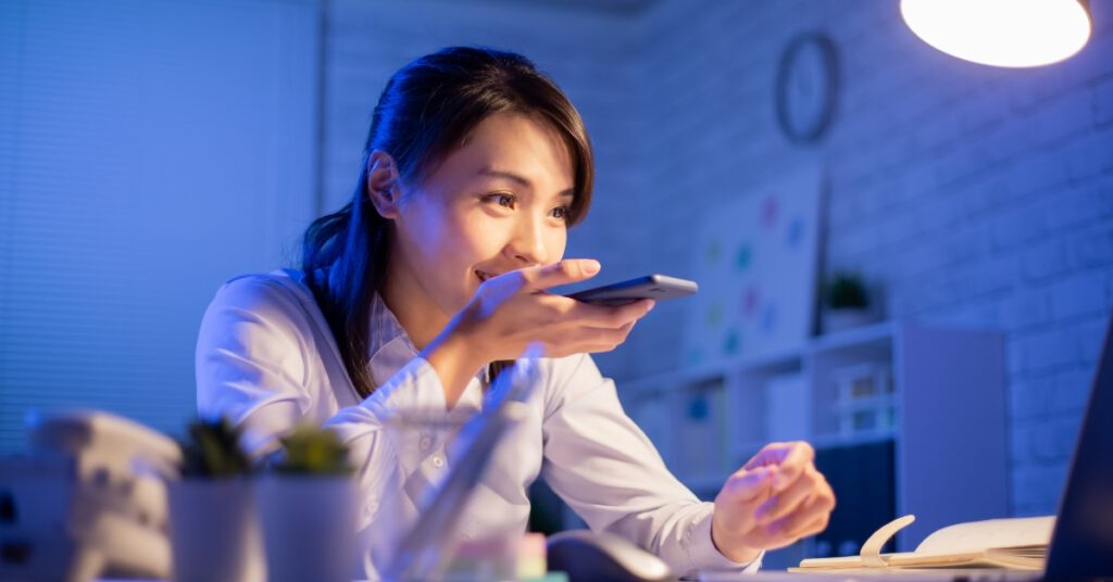 Asian woman sitting at a desk holding a mobile phone in front of a laptop using AI