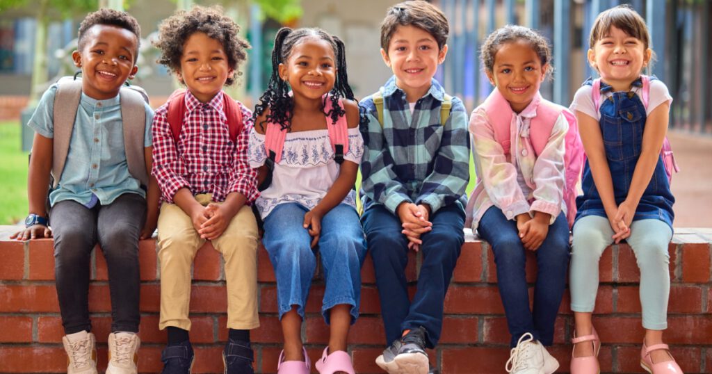 Portrait Of Multi-Cultural Elementary School Pupils Sitting On Wall Outdoors At School