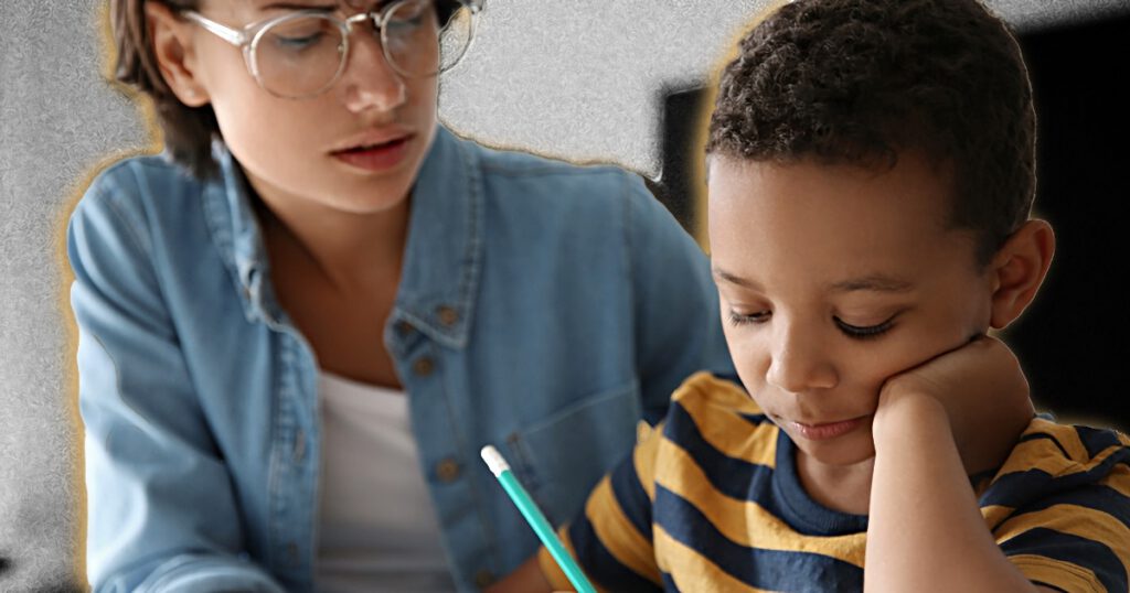White woman teacher sitting with a young Black boy in a classroom completing school work.