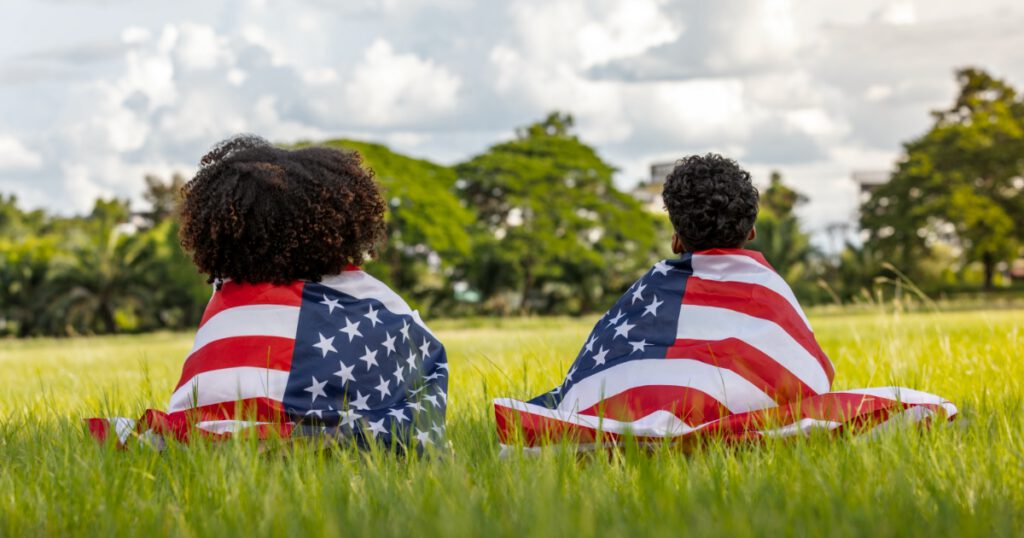 Two black children with textured hair, wrapped in American Flags, sitting in a field of grass