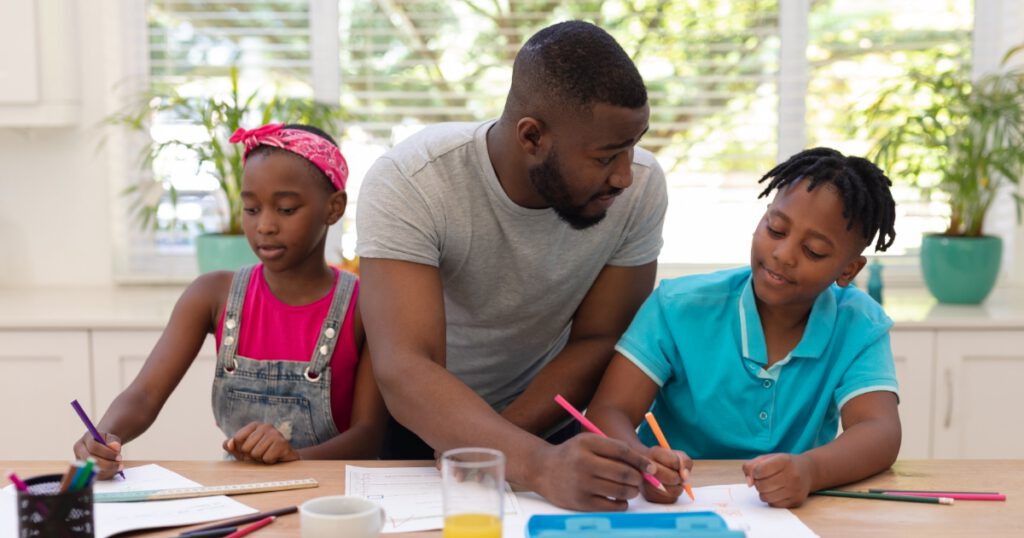 Black fathers sitting at a table with two Black children doing homework.