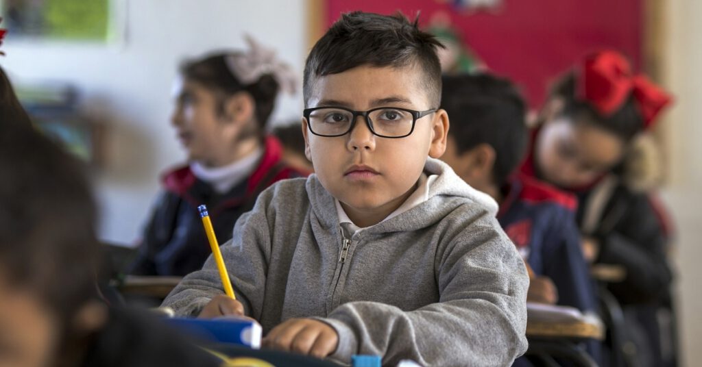 Young Latino boy sitting at a desk in a classroom wearing a sweatshirt and glasses and holding a pencil; looking at the camera