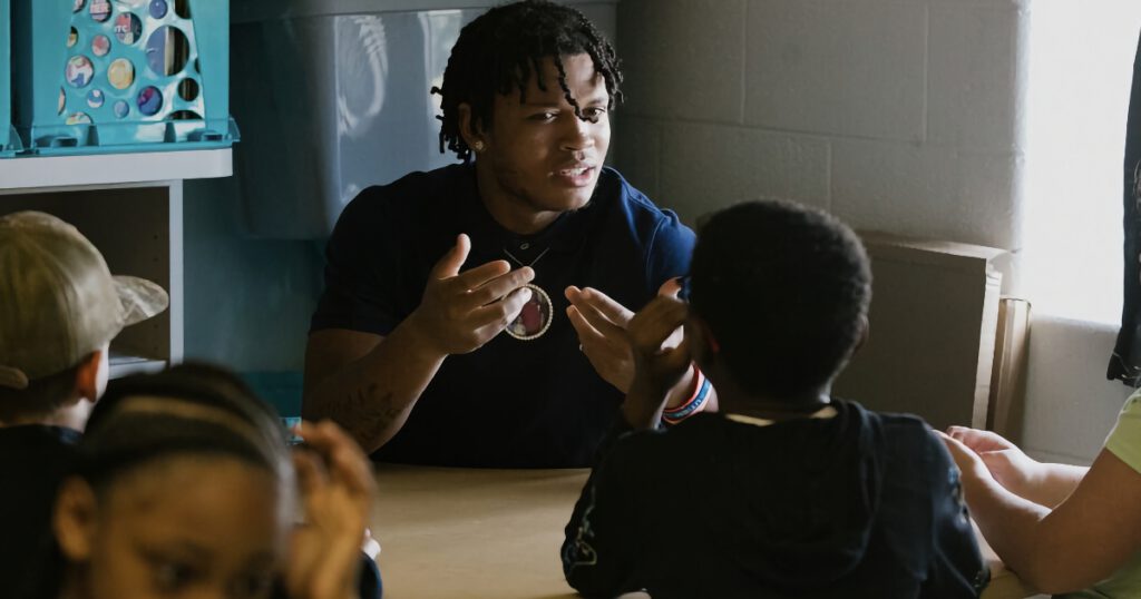 Young, Black, male teacher sitting at a table in a classroom teaching students.