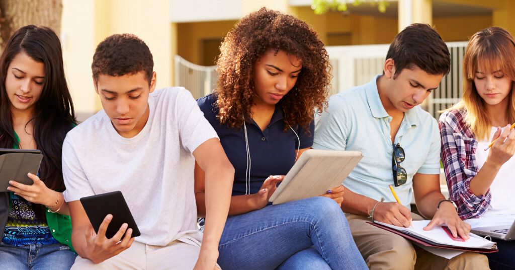 Diverse high school students woking on tablets and notebooks outdoors