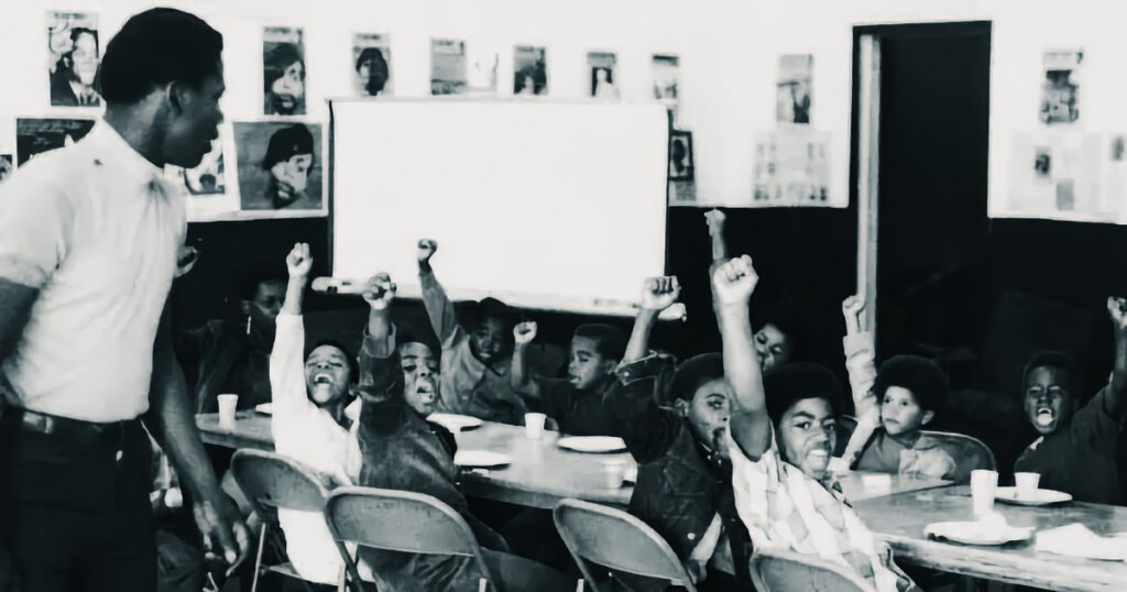 Member of the Black Panther Party in a school with a table full of Black boys giving a salute
