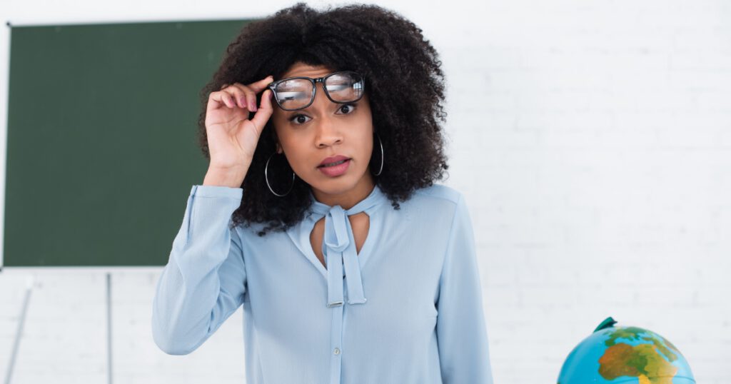 Shocked Black female educator looking out from underneath her glasses, standing in front of a chalk board.